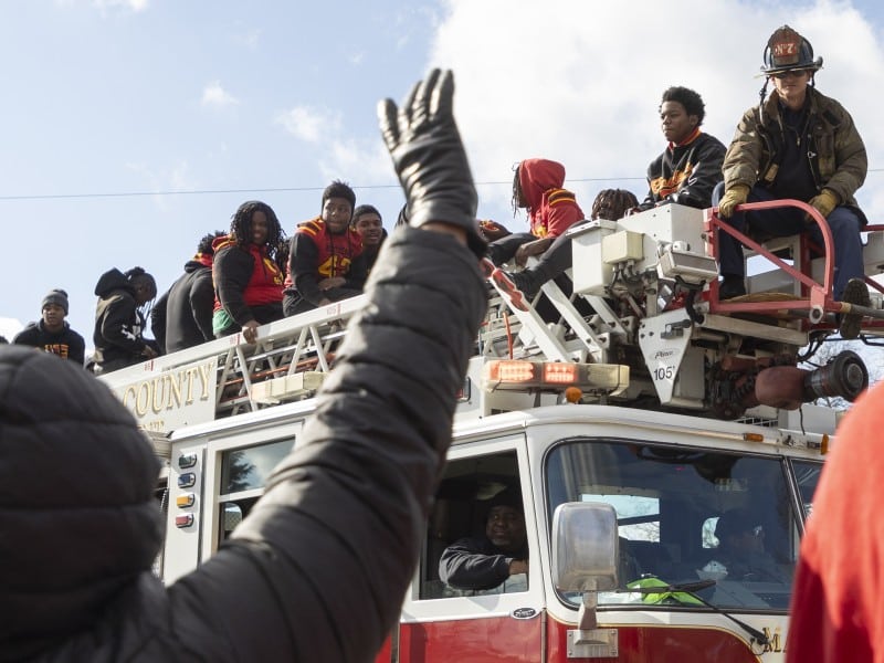A group of young people, some wearing red and yellow sports jerseys, sit on top of a fire truck's ladder, smiling and waving during an event or parade. A firefighter sits on the rear of the truck, and a driver is visible in the cab. In the foreground, a person raises a gloved hand toward the truck. The sky is bright and partly cloudy.