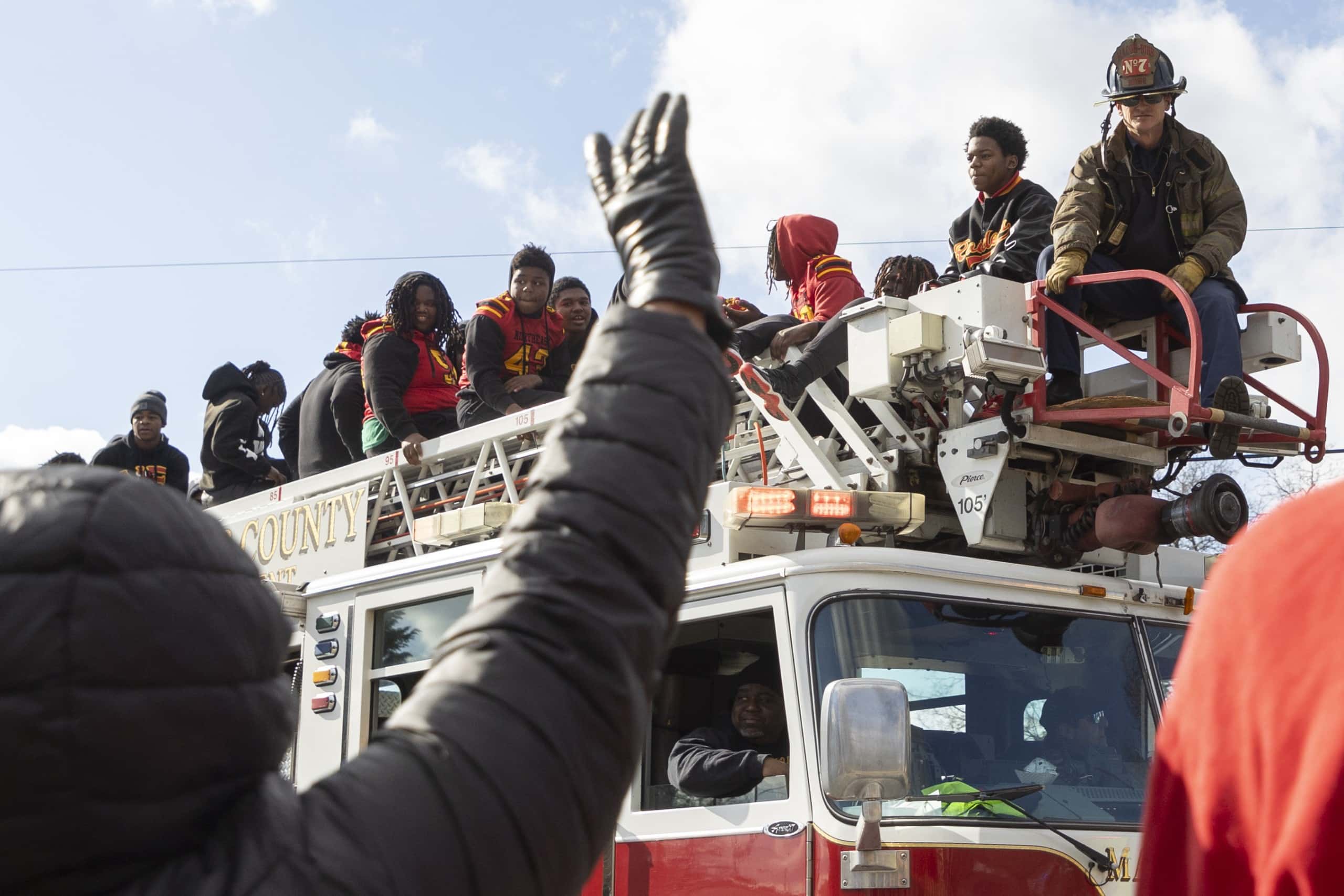 A group of young people, some wearing red and yellow sports jerseys, sit on top of a fire truck's ladder, smiling and waving during an event or parade. A firefighter sits on the rear of the truck, and a driver is visible in the cab. In the foreground, a person raises a gloved hand toward the truck. The sky is bright and partly cloudy.