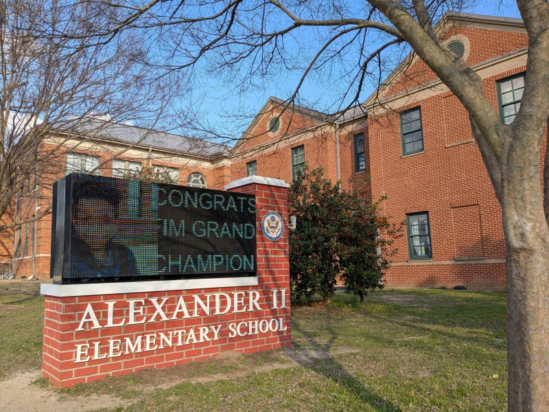The image shows the exterior of Alexander II Elementary School, a red brick building with multiple windows and a gray roof. In the foreground, a brick sign with white lettering displays the school's name.