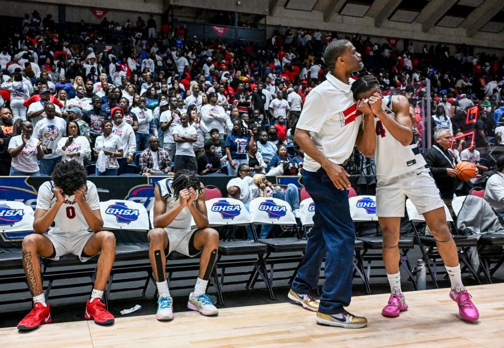 A basketball game at Macon Coliseum with the crowd cheering, with many fans standing, raising their arms, and celebrating. Players on the court, wearing white and black uniforms, also react to the moment. The large arena is packed with spectators, and the excitement is visible throughout the scene.