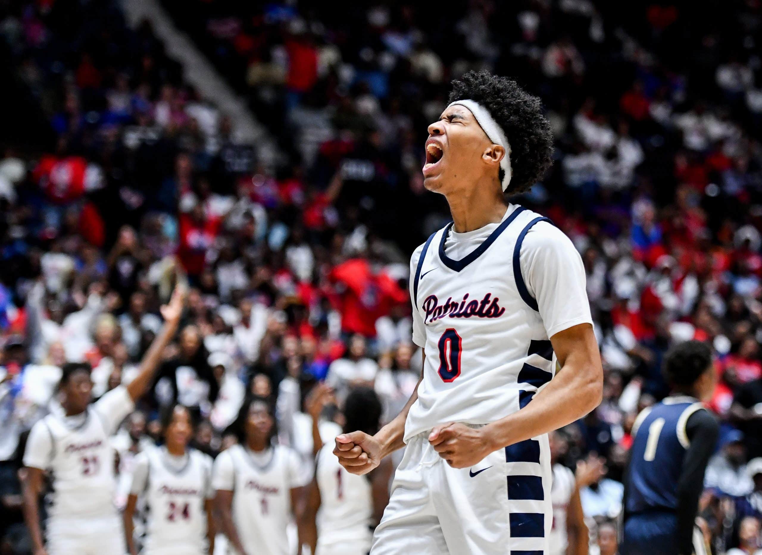 A basketball player wearing a white "Patriots" jersey with the number 0 shouts in celebration, clenching his fists. He has a white headband and curly hair, and his face shows intense emotion. Behind him, teammates in matching white uniforms and a crowd of fans, many dressed in red, cheer with raised arms. An opposing player in a dark uniform stands in the background.